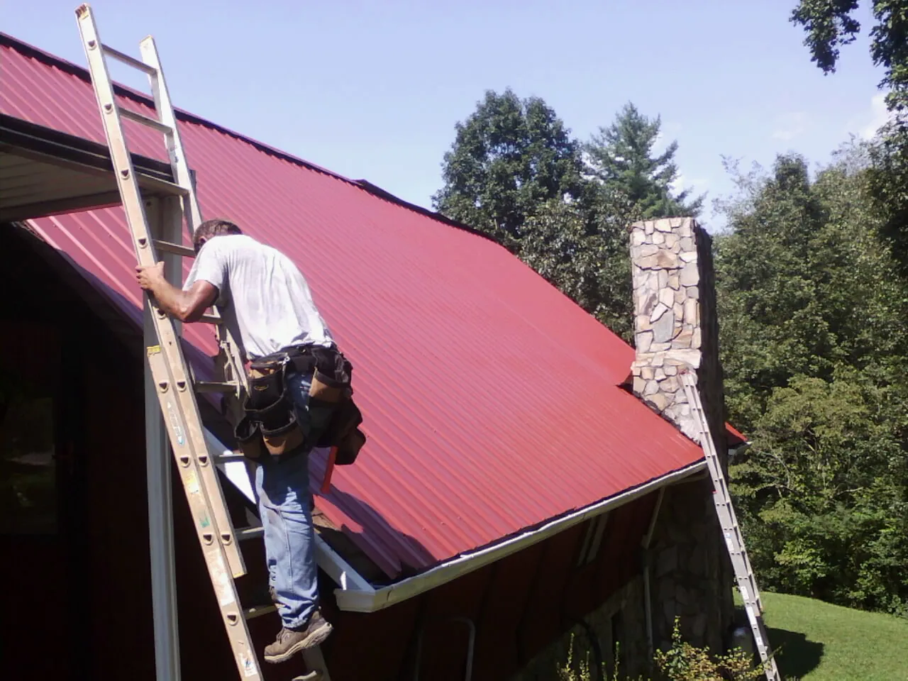 Roofer on a ladder beside a red metal roof and stone chimney — Soffit & Fascia Repair by Roofing Brothers in West Suffield